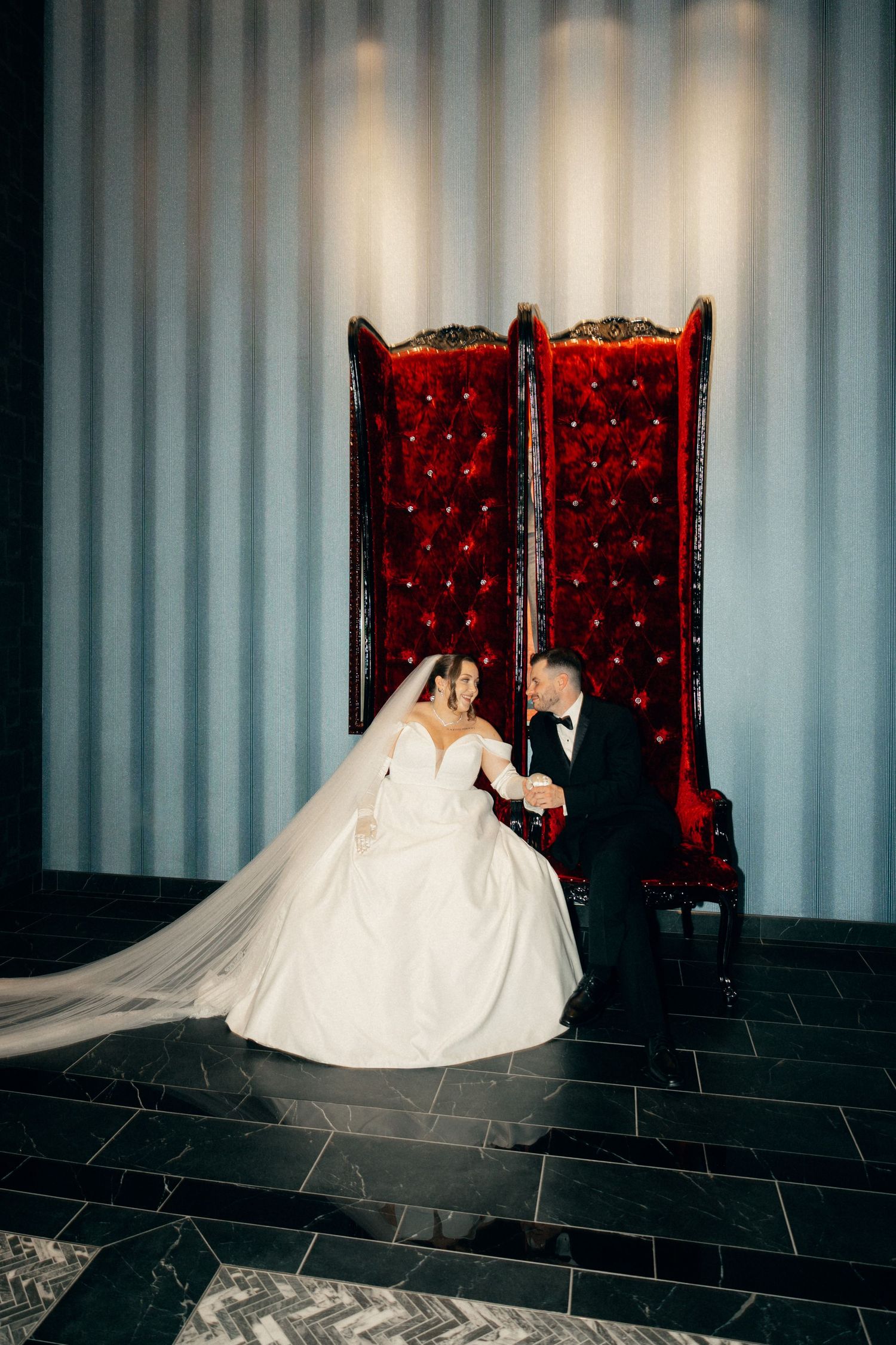 Romantic wedding portrait series against red velvet chairs and white curtain backdrop.