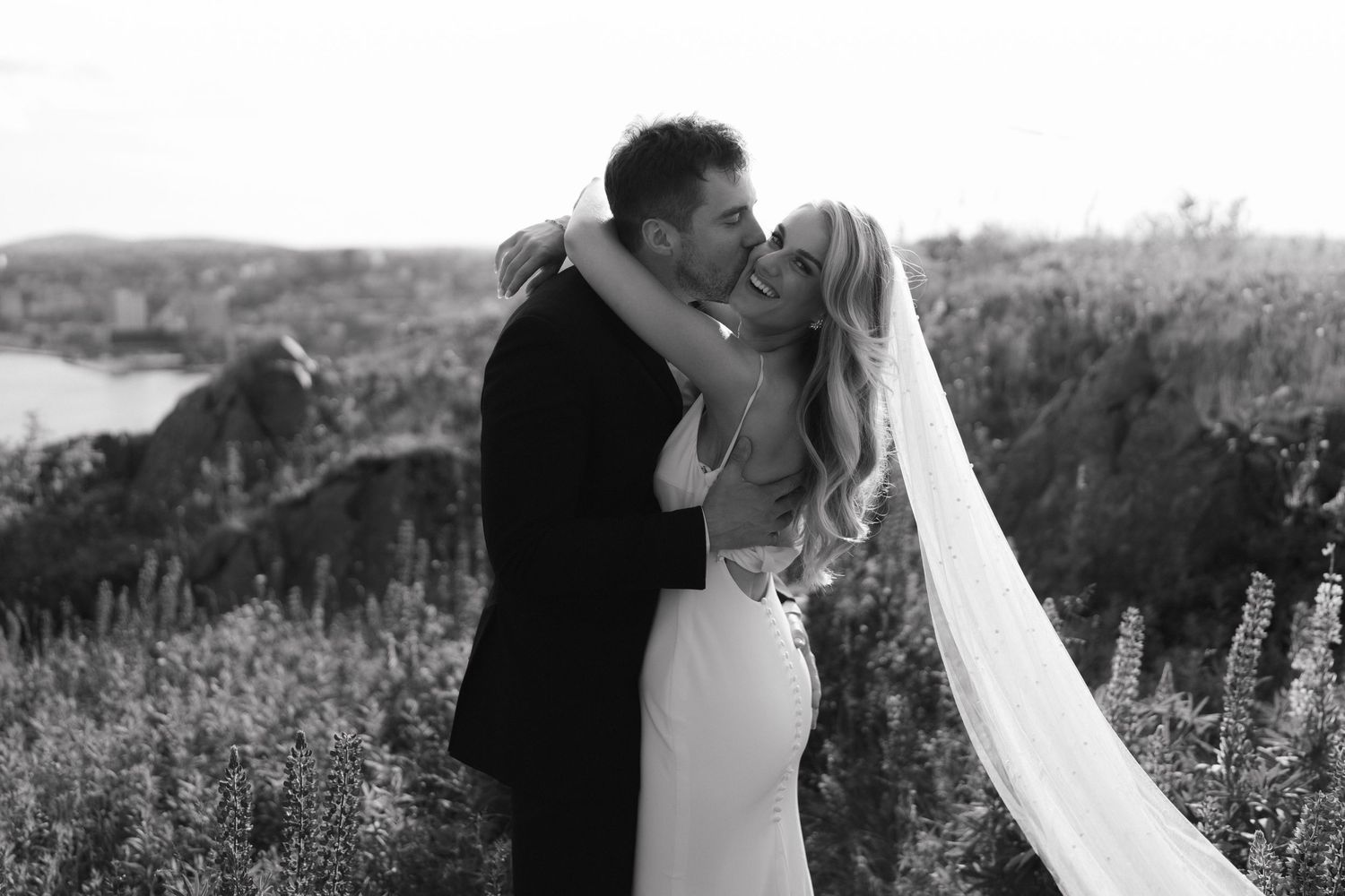 A romantic wedding couple shares a tender embrace against a coastal backdrop with flowing veil and wildflowers.