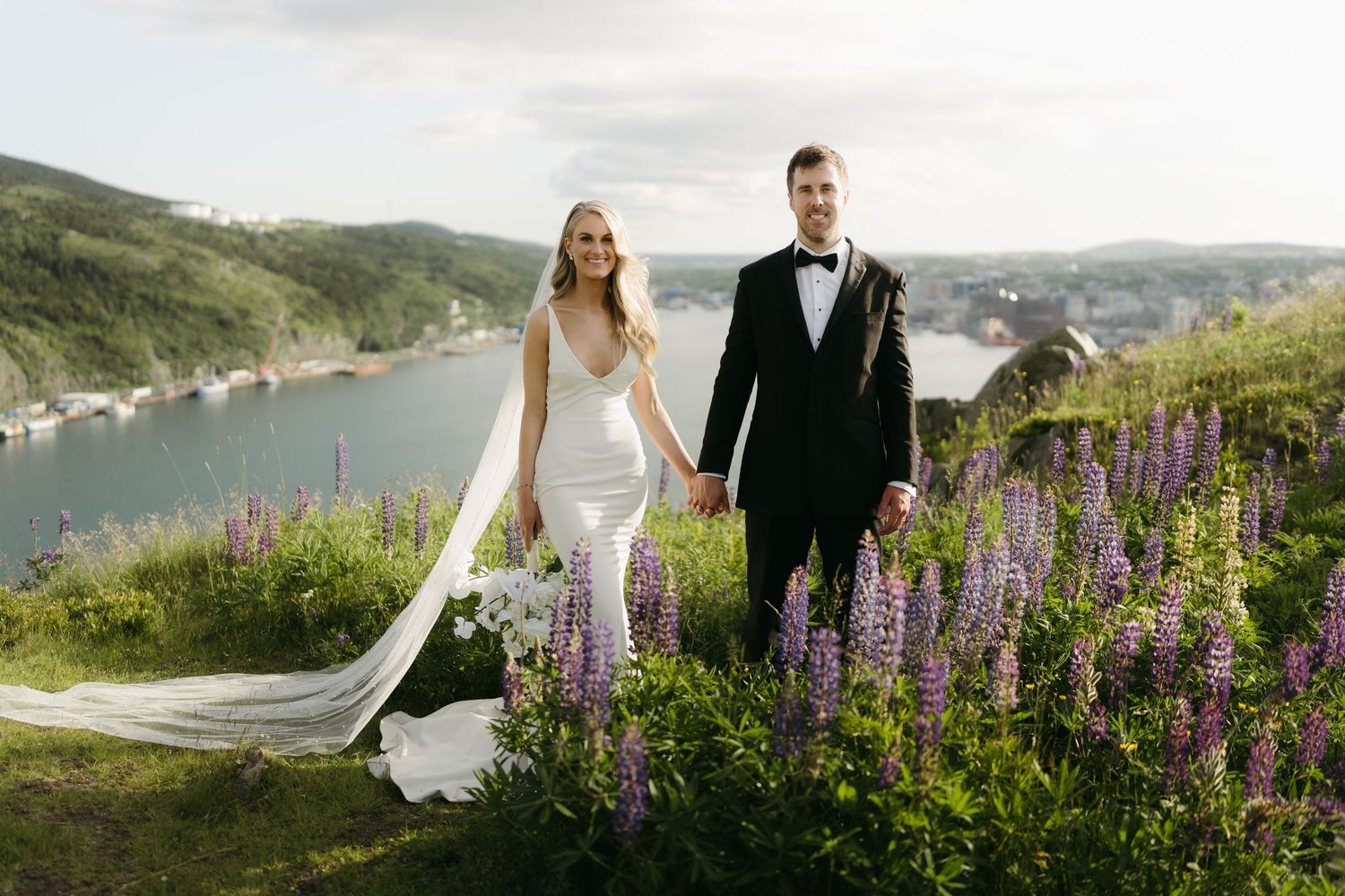 Newlyweds stroll together along a coastal path with purple wildflowers in bloom.