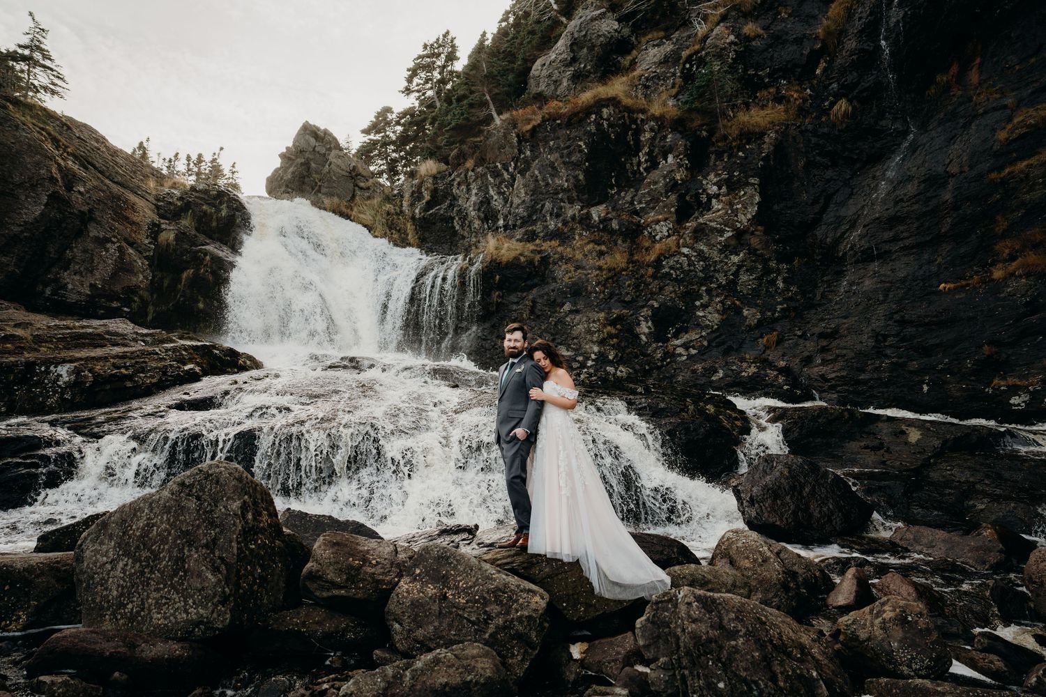 A picturesque waterfall serves as backdrop for an intimate wedding moment on rocky terrain.