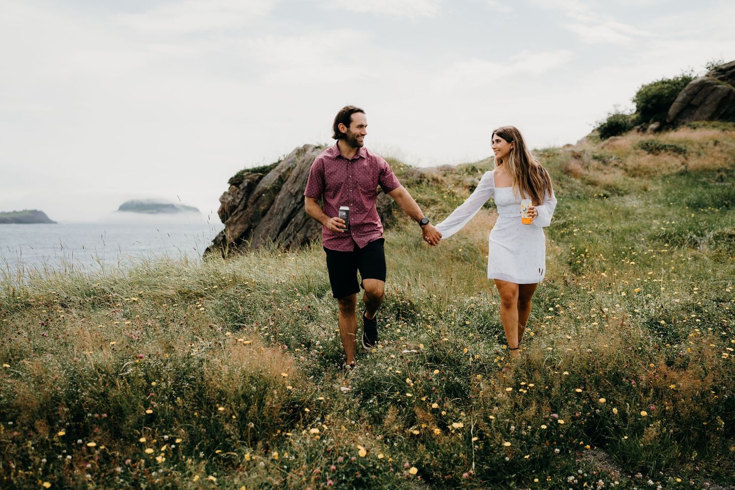 Couple walks hand in hand through wildflower field by the ocean.