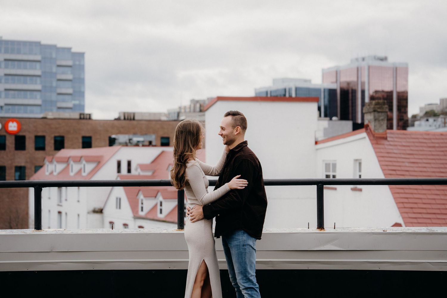 Two people share an intimate moment on a rooftop terrace overlooking brick buildings and modern architecture.