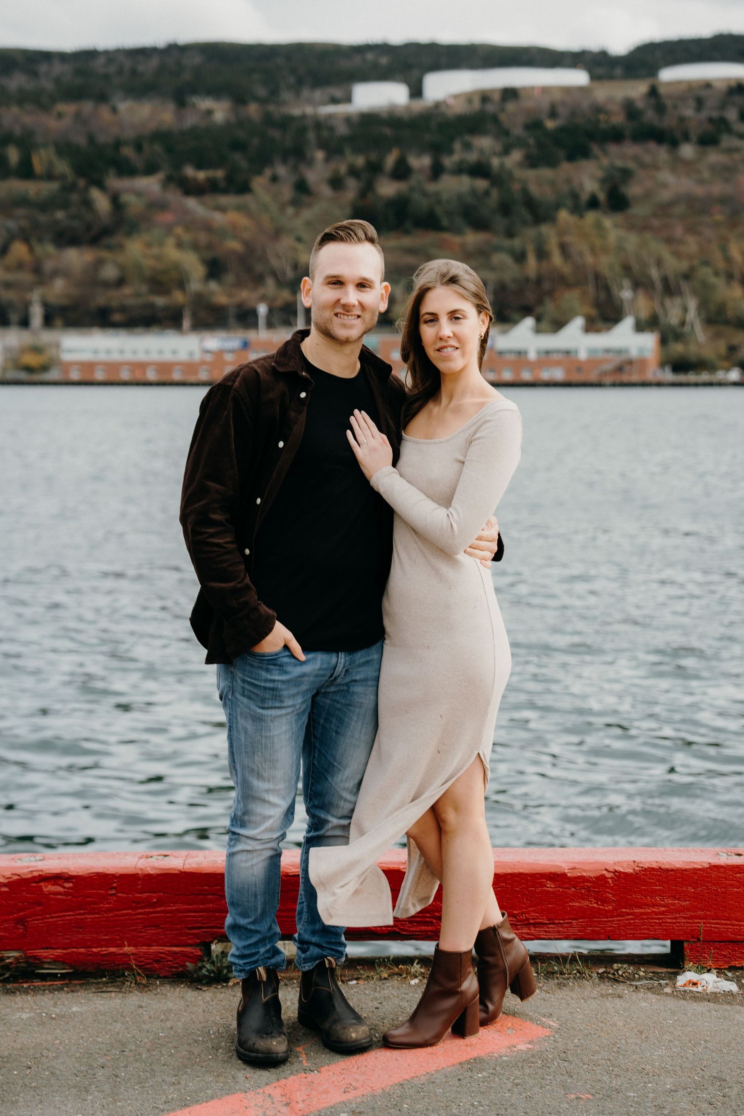 Couple posing by waterfront with autumn hillside and industrial buildings in background.