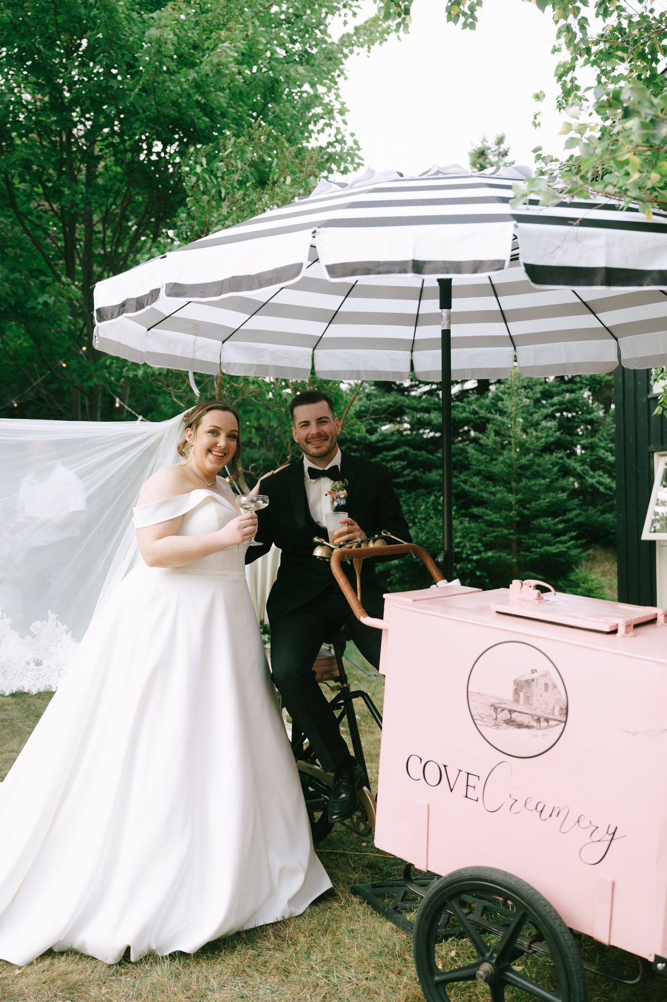 Wedding celebration featuring a pink and white ice cream cart with striped umbrellas in a garden setting.