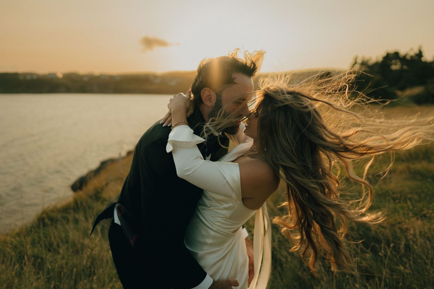 A romantic couple embraces on a grassy hillside overlooking a lake at sunset as her hair flows in the wind.