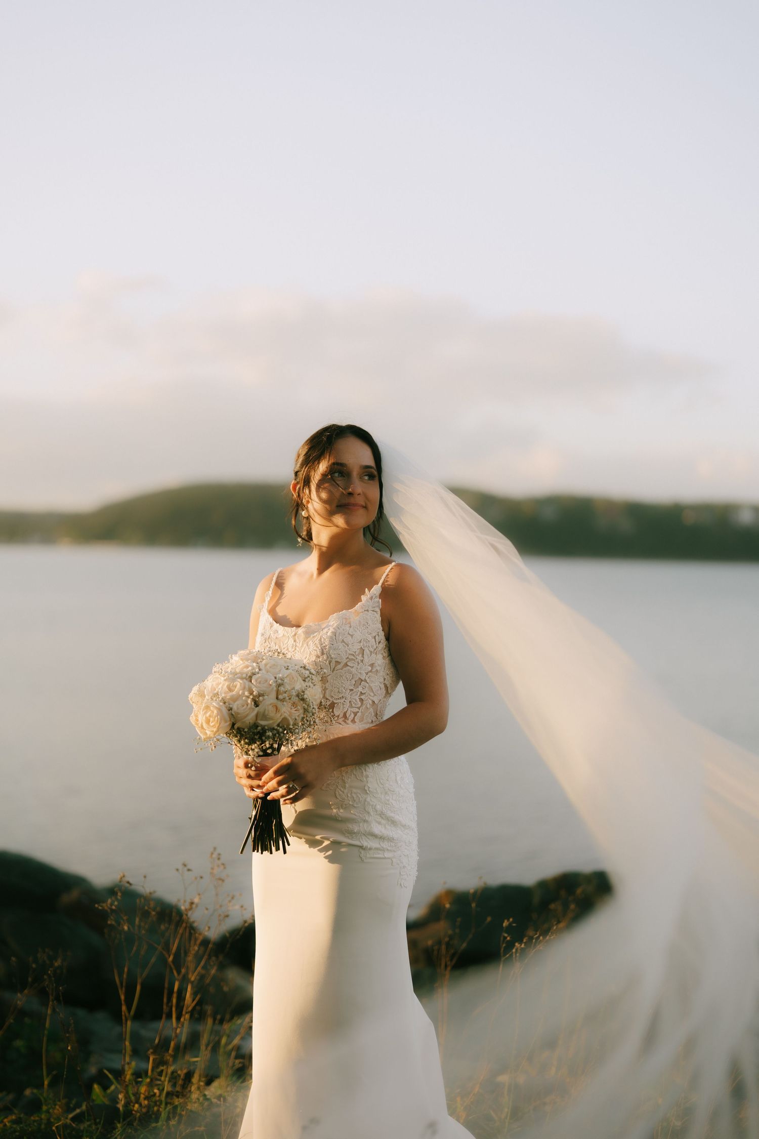 Bride in flowing white wedding dress with veil stands against scenic mountain landscape at sunset.