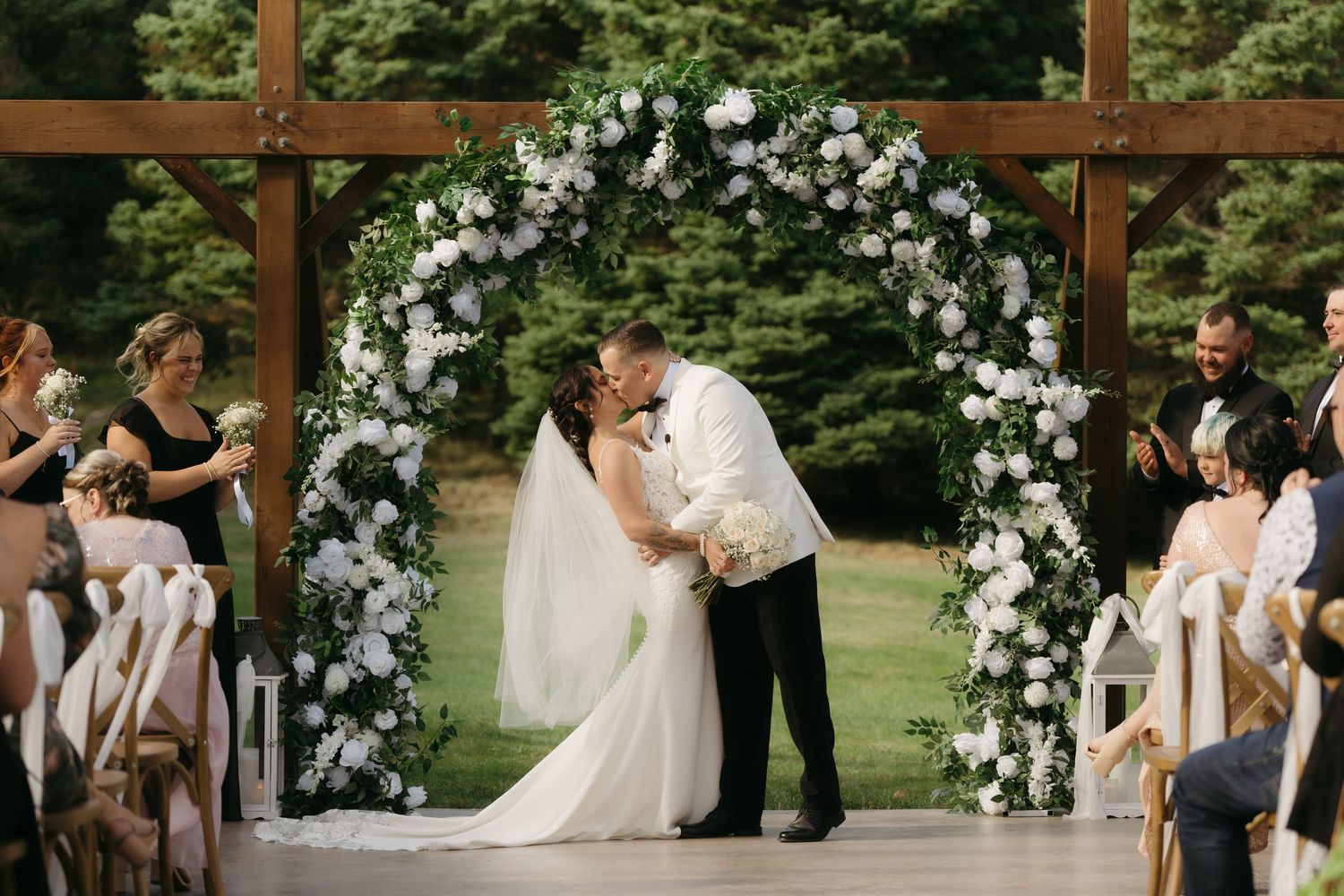 A wedding ceremony takes place outdoors under a wooden pergola adorned with white floral arrangements.