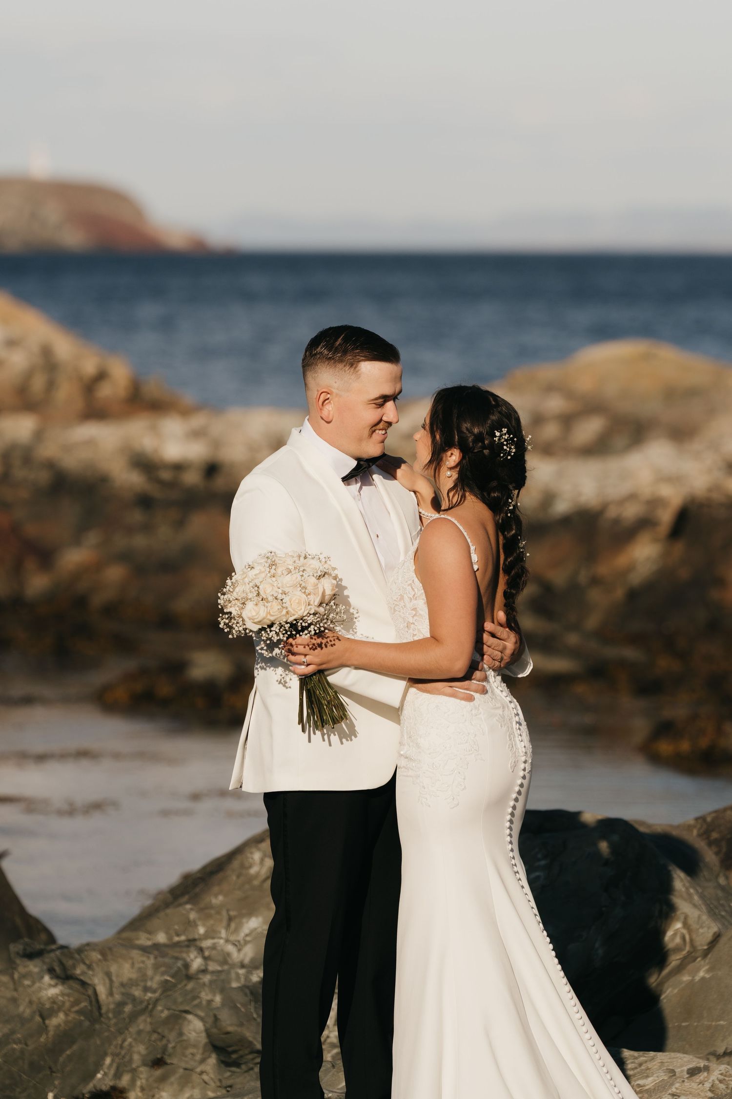 A romantic wedding couple embraces on rocky beach at sunset with ocean and bouquet of baby's breath flowers.