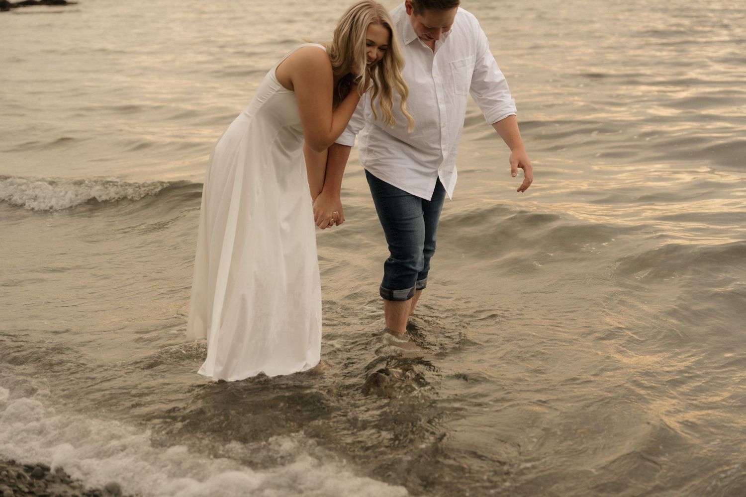 Couple walking together in shallow ocean water at sunset with their white flowing dress and casual attire.
