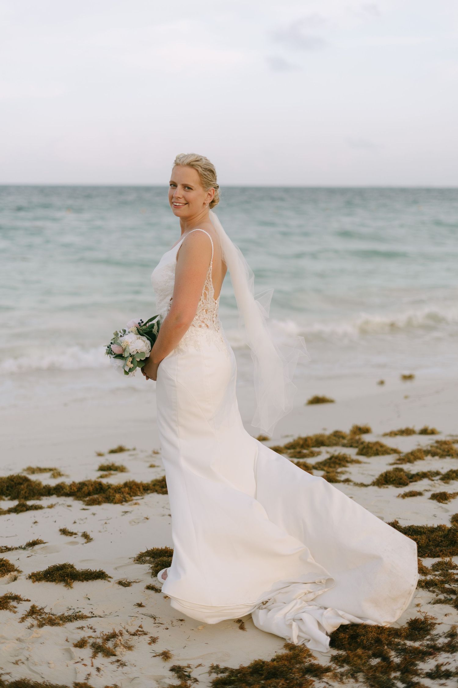 Bride in flowing white dress twirling on beach with ocean view.