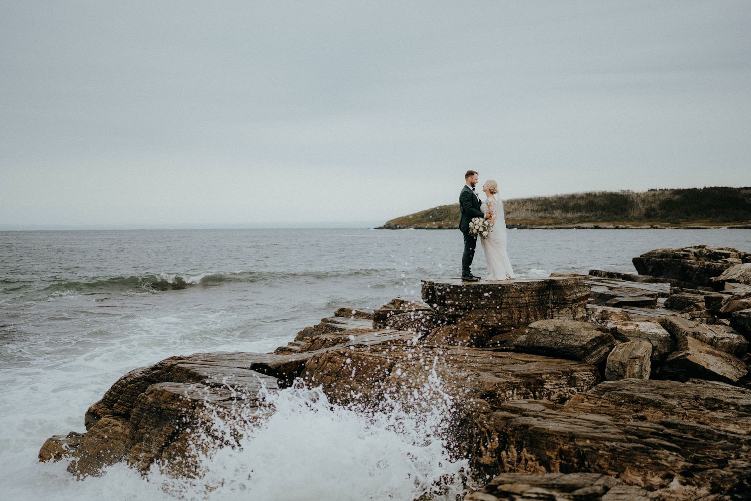 A couple standing on rocky shore with waves crashing and ocean views.