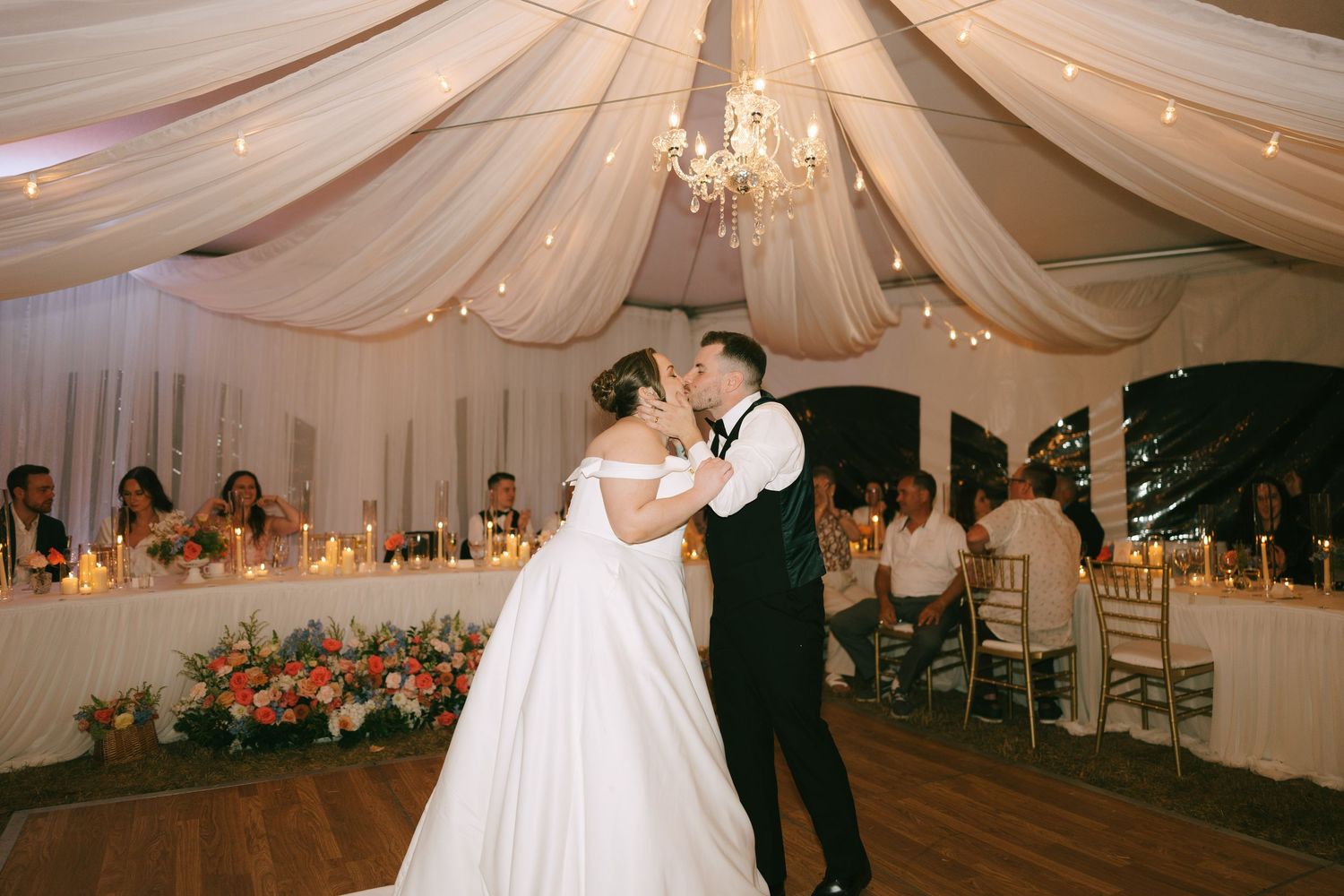 Wedding guests watch as a couple dances beneath flowing white fabric and crystal chandeliers.
