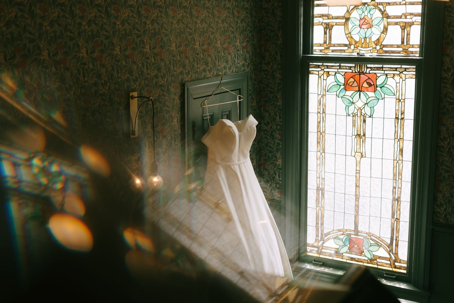 Stained glass window casting colorful light on hanging white wedding dress.
