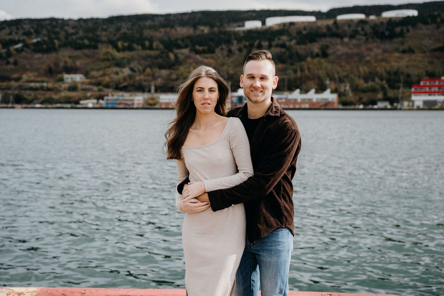 A loving embrace captured against a coastal landscape with industrial buildings in the background.