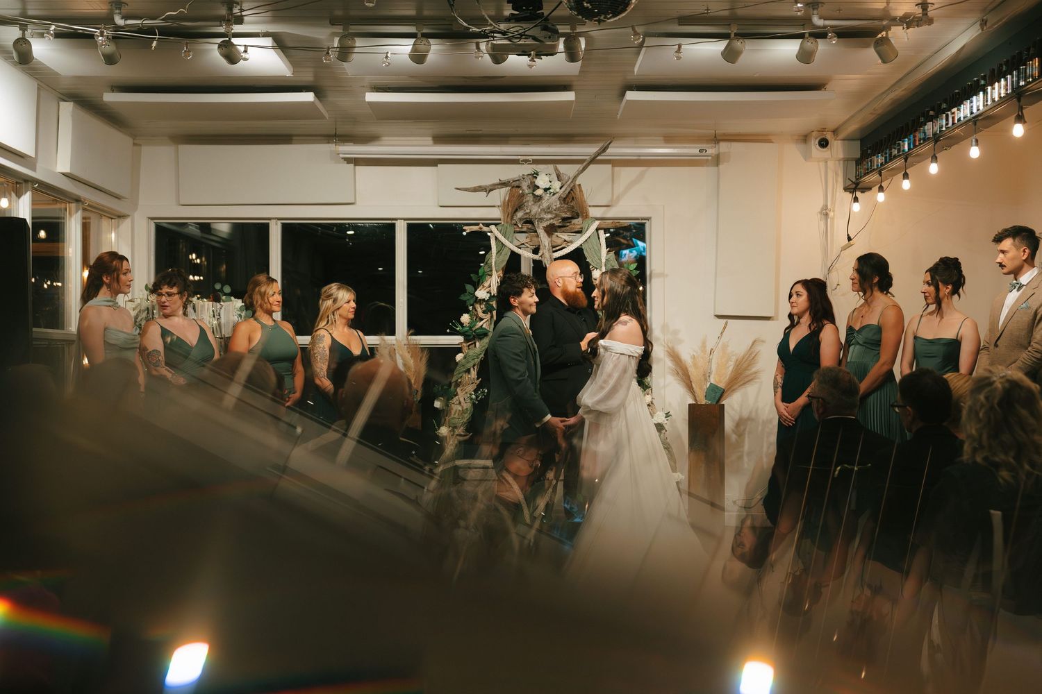 A crowd of people gather in a dimly lit art gallery space with track lighting overhead and exposed pipes on the ceiling.