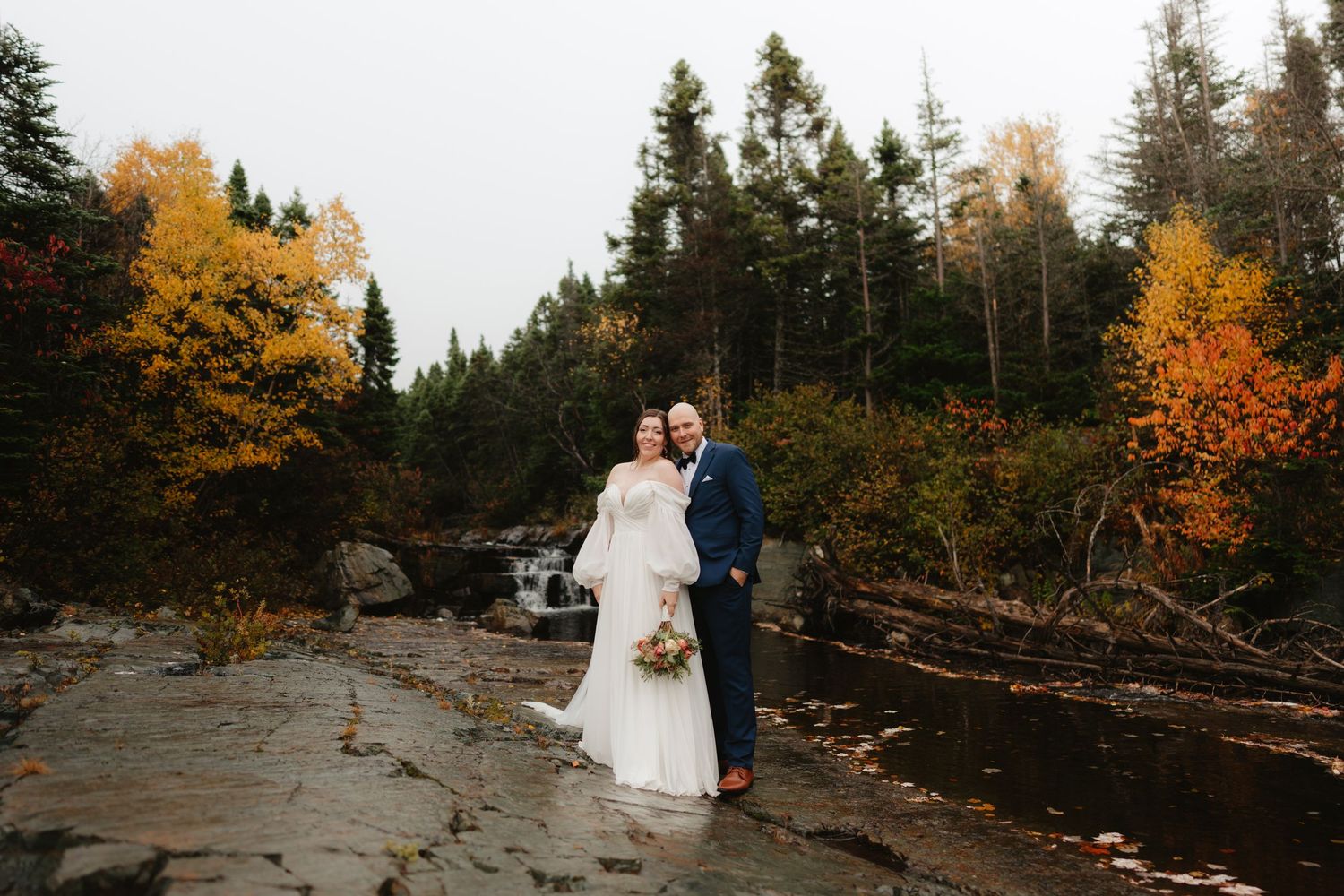 A couple poses for wedding photos in a scenic autumn forest setting with vibrant orange foliage and fallen logs.