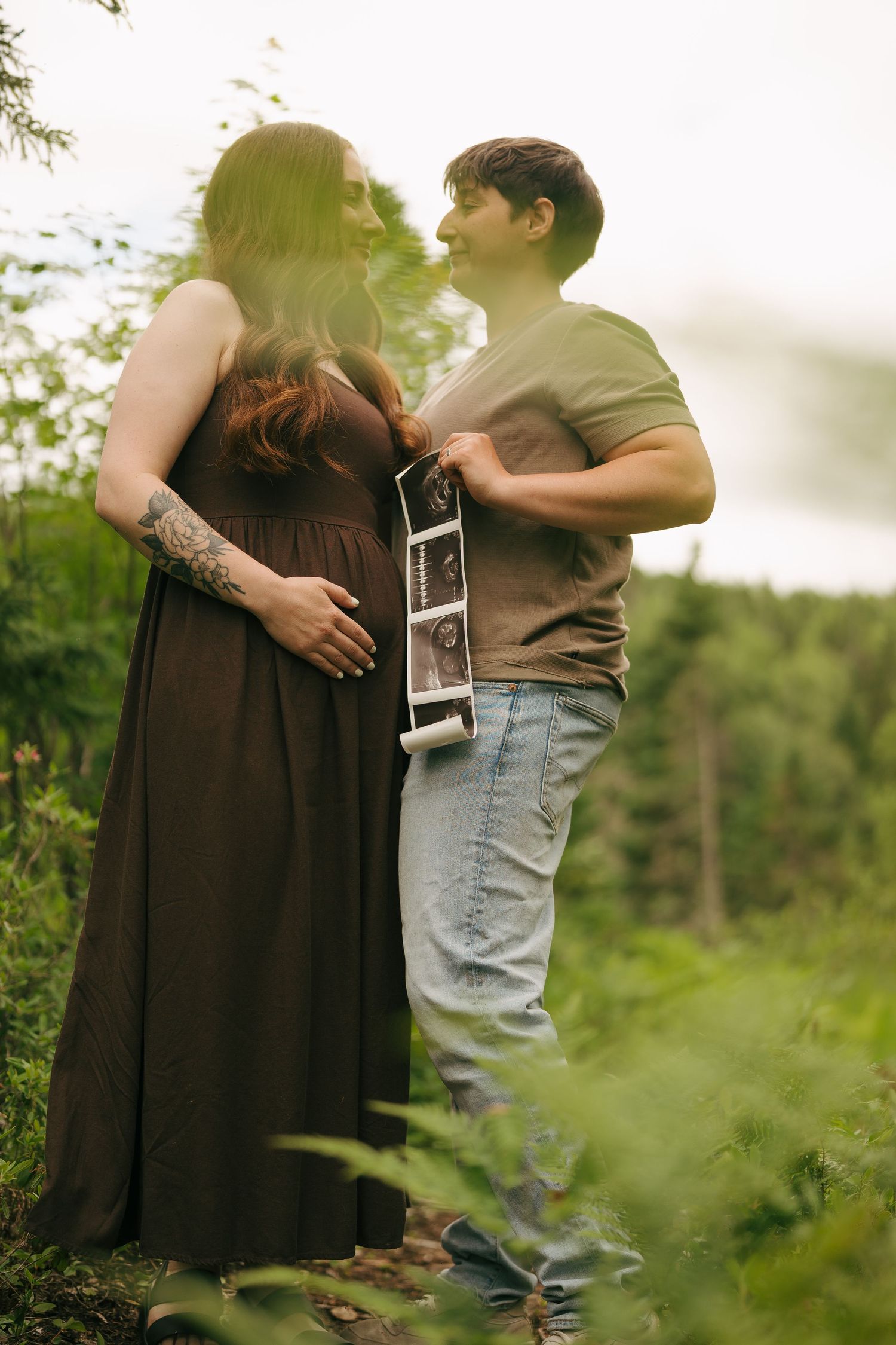 Pregnant couple embracing outdoors in lush greenery, man holding ultrasound photos, woman in brown dress with tattoo.
