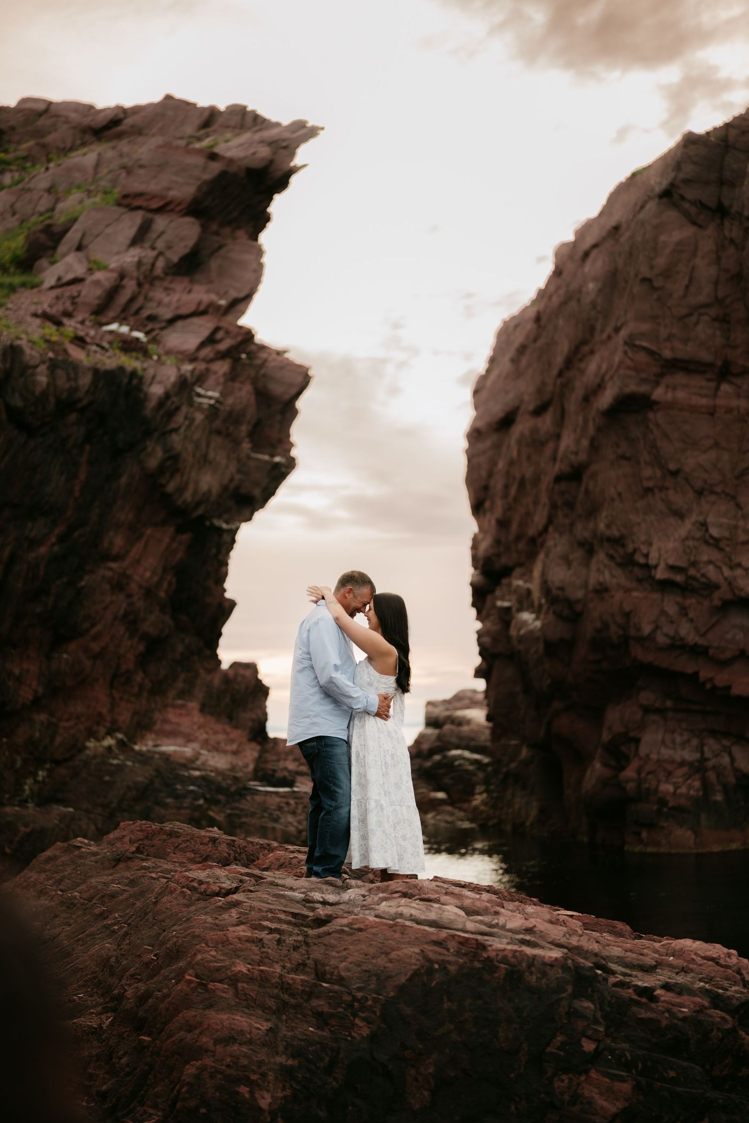Couple kissing between dramatic red rock formations on a rocky coastline at sunset during engagement photo session.