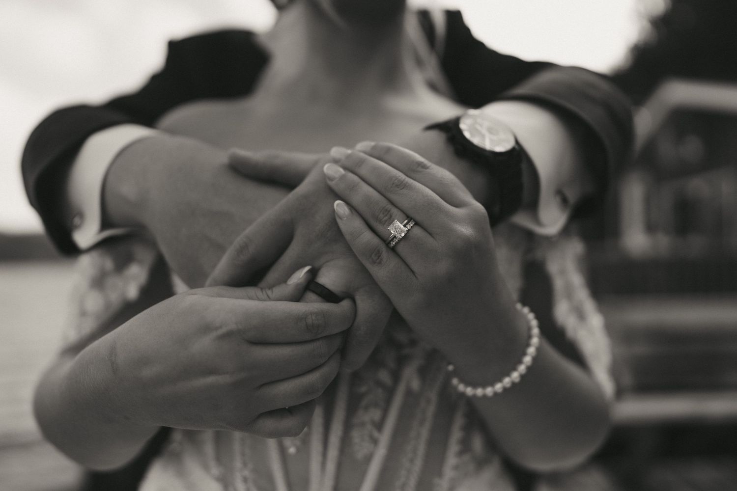 Black and white photo of couple holding hands showing engagement ring and pearl bracelet at waterside wedding.