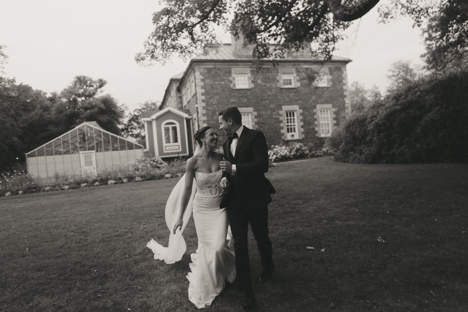 Black and white photo of a bride and groom walking on a lawn in front of a historic stone mansion on their wedding day.