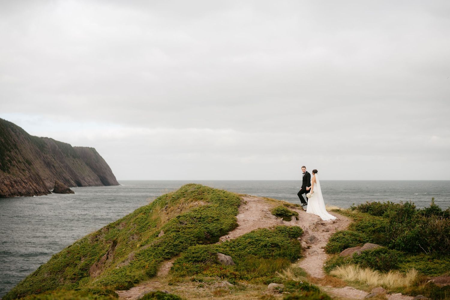 Bride and groom stand on a scenic coastal cliff overlooking the ocean on their wedding day.