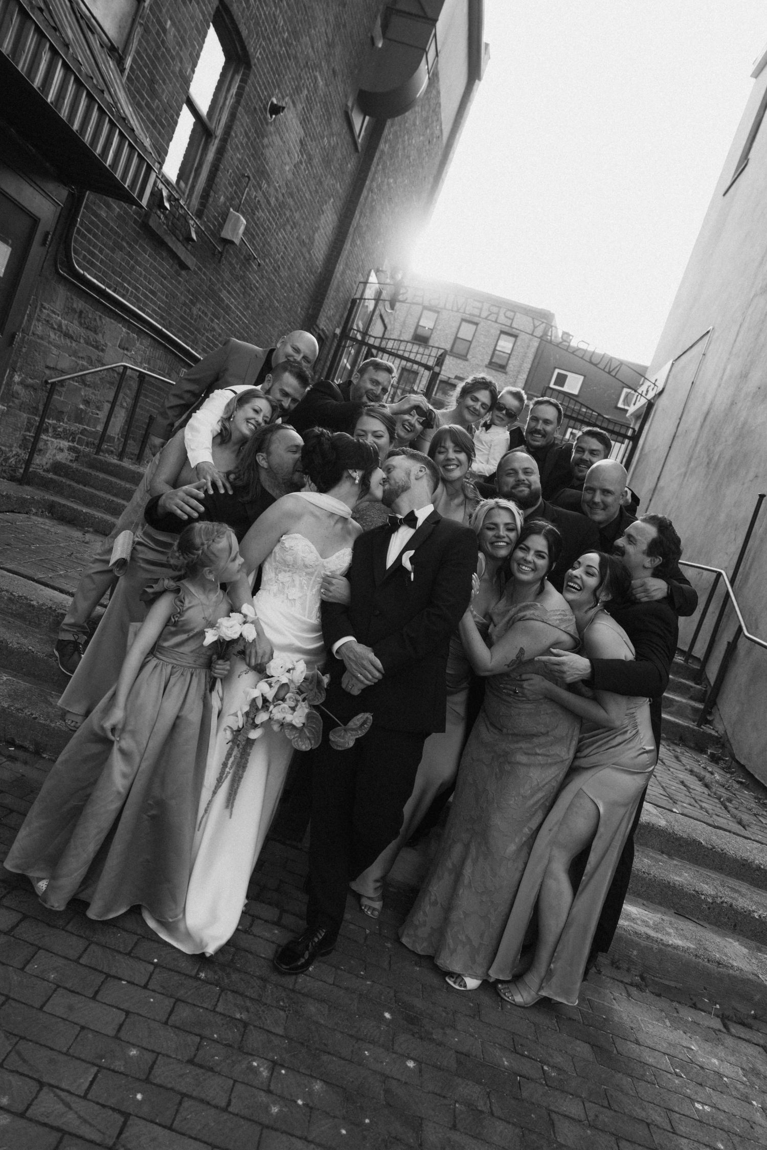 Black and white wedding photo of bride and groom kissing surrounded by joyful wedding party in urban alley.