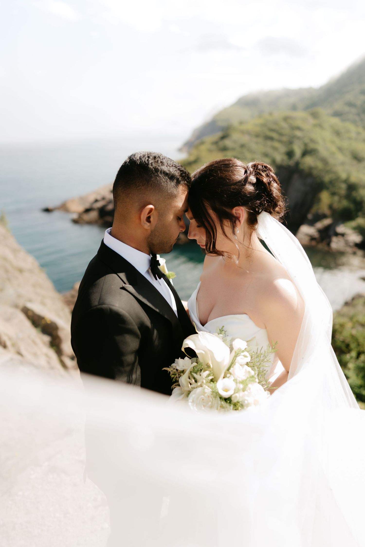 Bride and groom share romantic moment on coastal cliff, bride holding white bouquet with ocean and green hills behind.