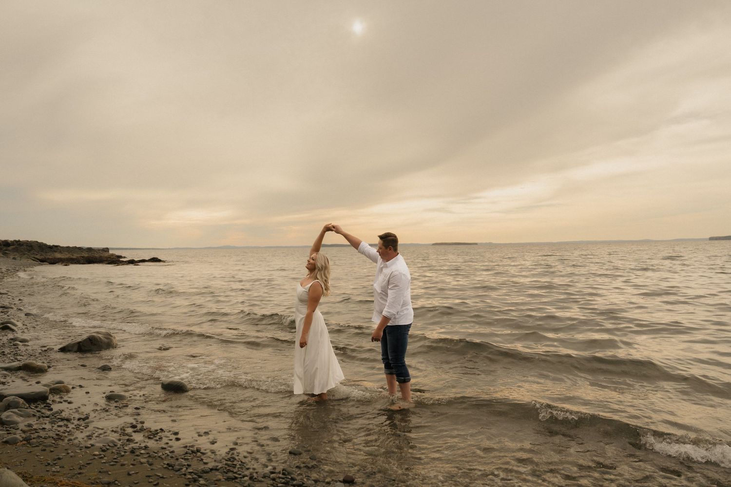 Couple in wedding attire walking along a rocky beach at sunset with dramatic cloudy sky over the ocean.