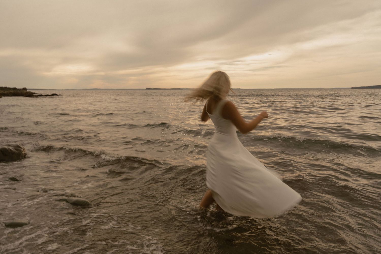 Ethereal figure in a flowing white dress walks along a misty beach shoreline at sunset.