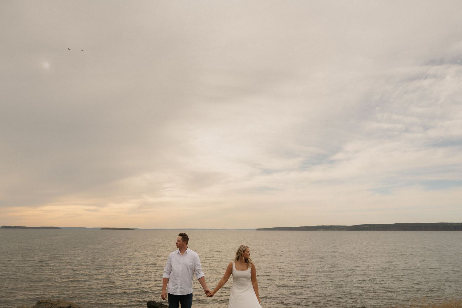 Couple holding hands while standing at the edge of a serene lake during sunset with cloudy skies.