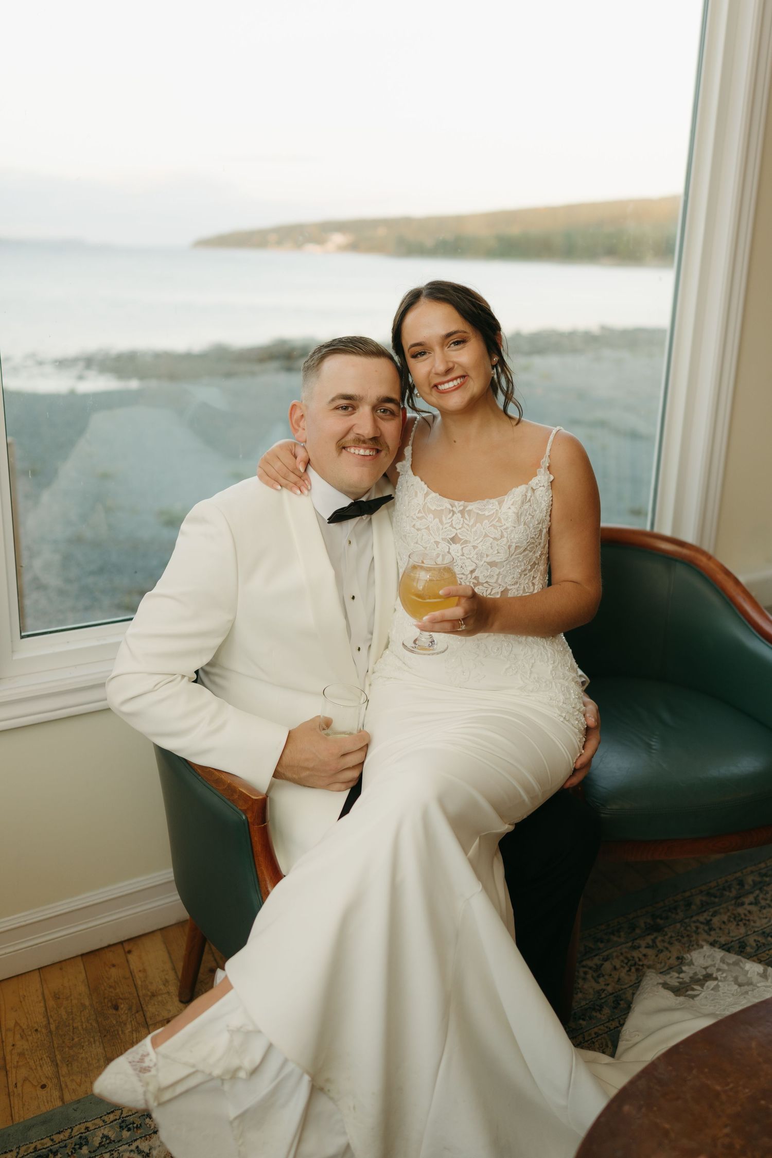 A couple in white wedding attire share intimate moments together while seated by large windows overlooking water.