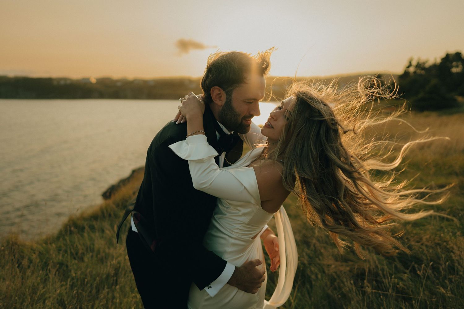 A couple embraces on a hilltop overlooking a lake during sunset as wind blows through their hair.