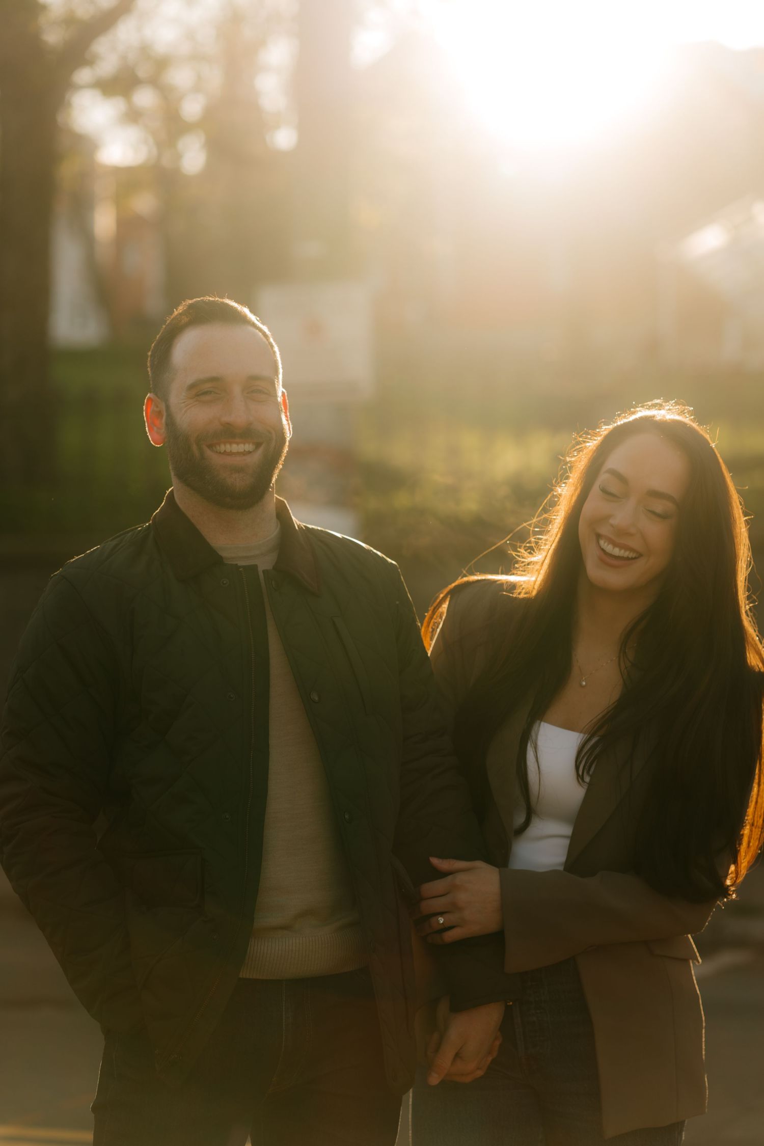 Smiling couple walking outdoors in golden sunset light, man with beard in dark jacket, woman with long dark hair laughing.