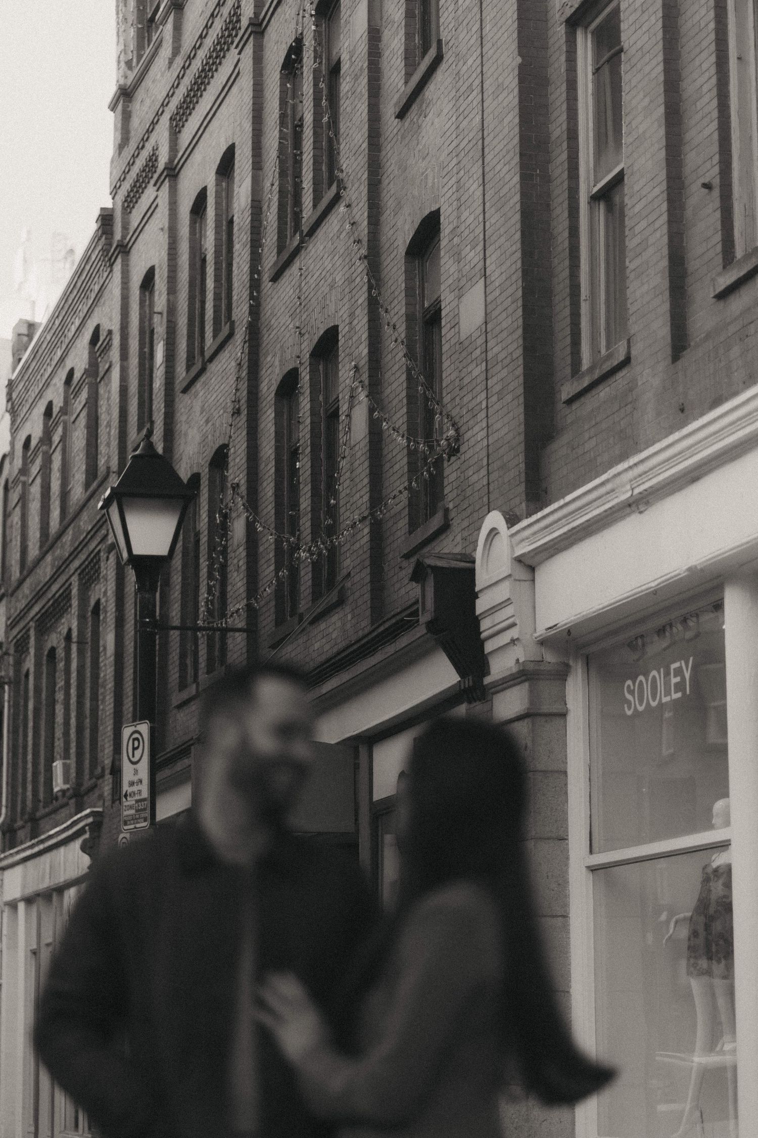 Black and white photo of a couple embracing on a cobblestone street with brick buildings and a vintage lamp post.
