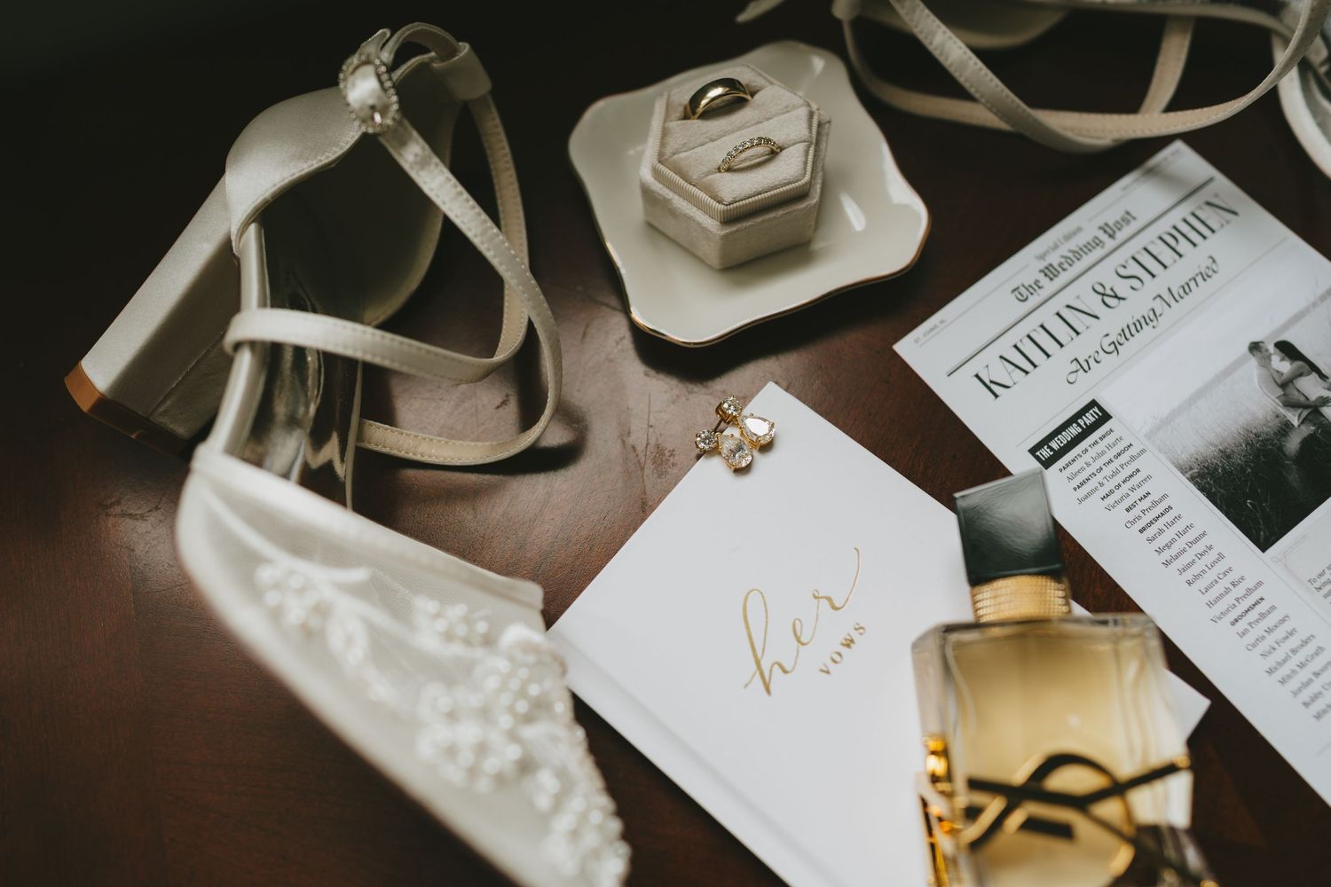 Elegant bridal flat lay featuring lace heels, gold ring box, pearl earrings, YSL perfume, and wedding program on dark wood.