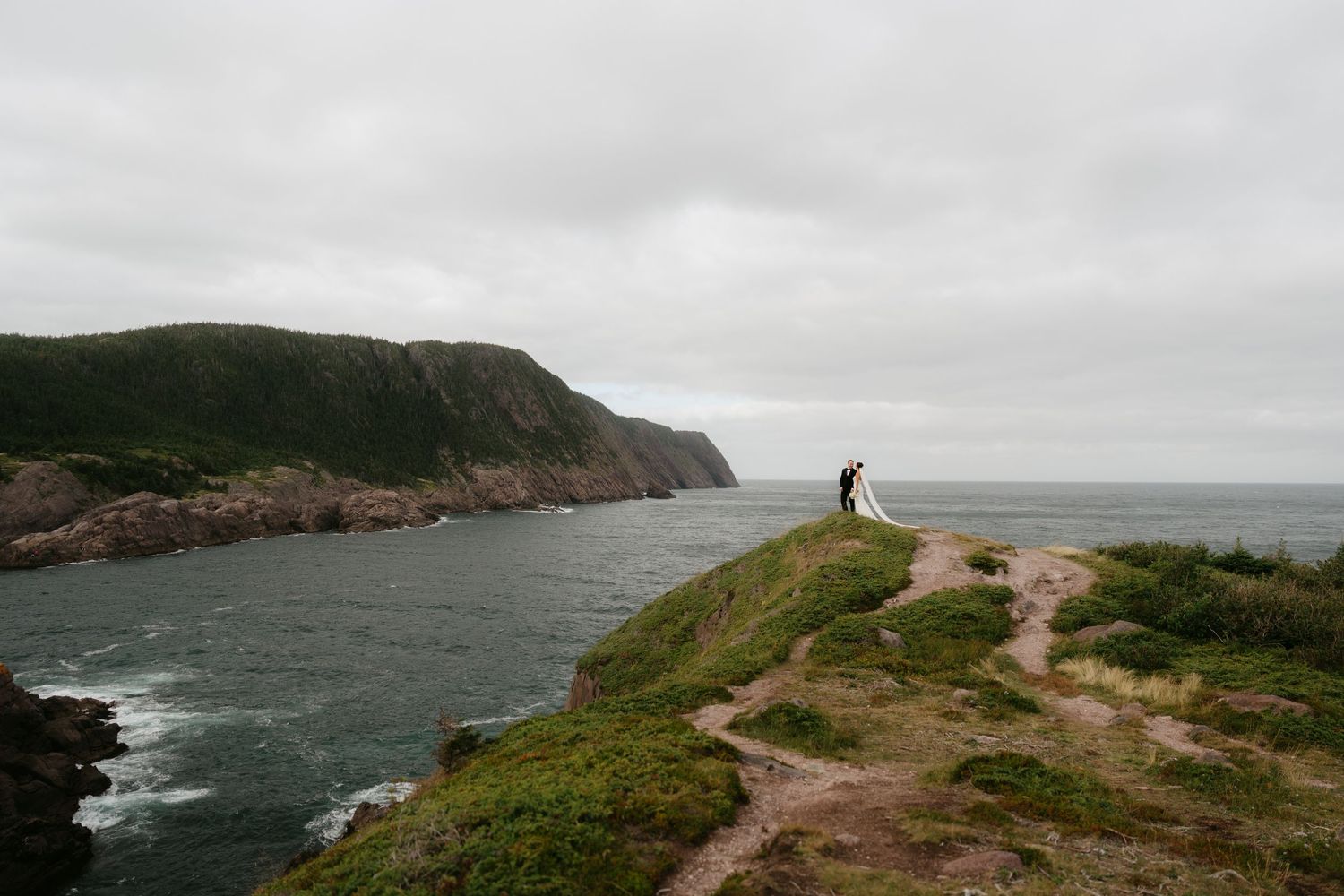 A lone hiker stands on a rugged coastal cliff overlooking a dramatic green headland and choppy ocean on an overcast day.