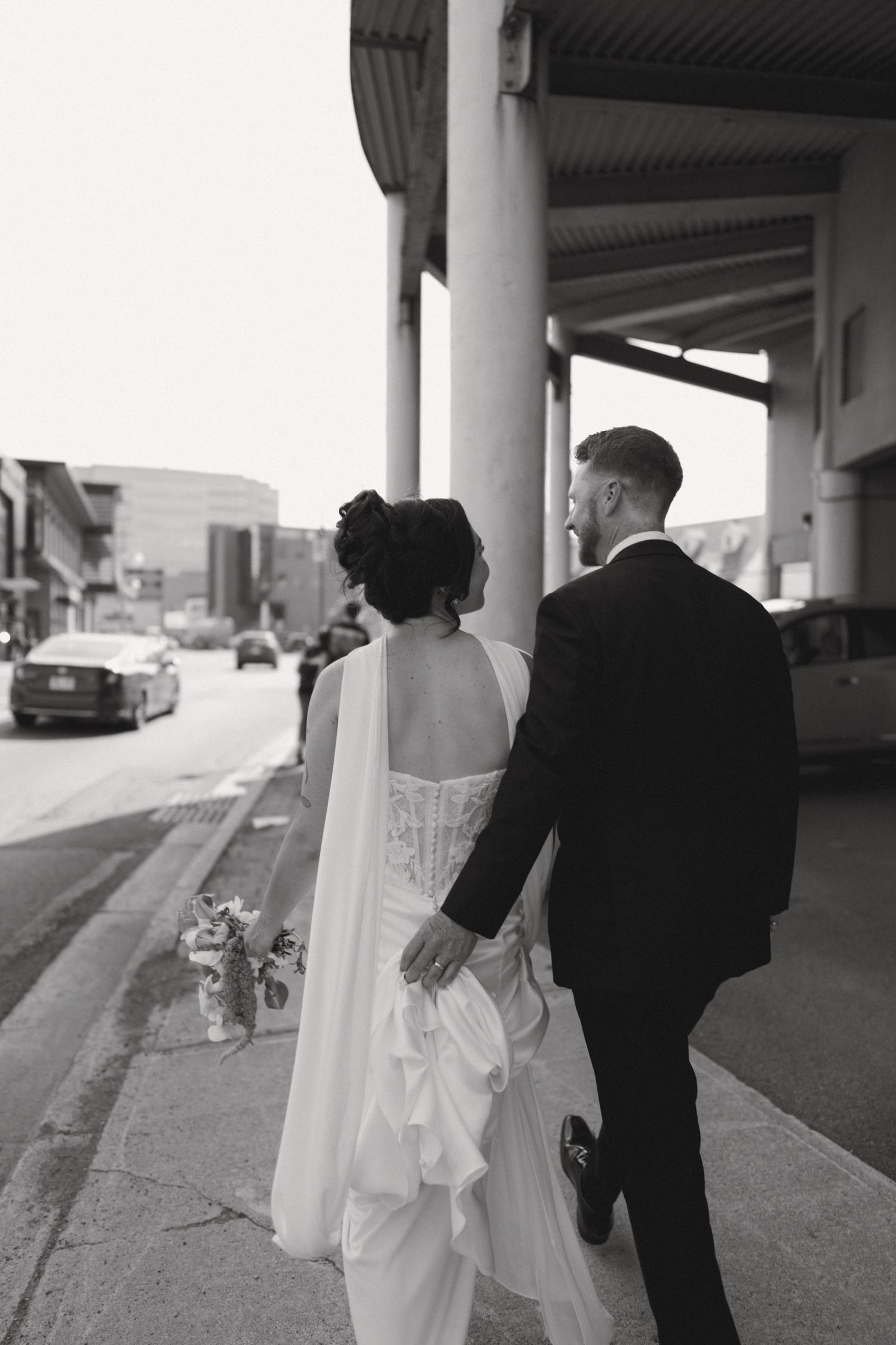 Black and white photo of a bride and groom walking hand in hand down a city sidewalk on their wedding day.