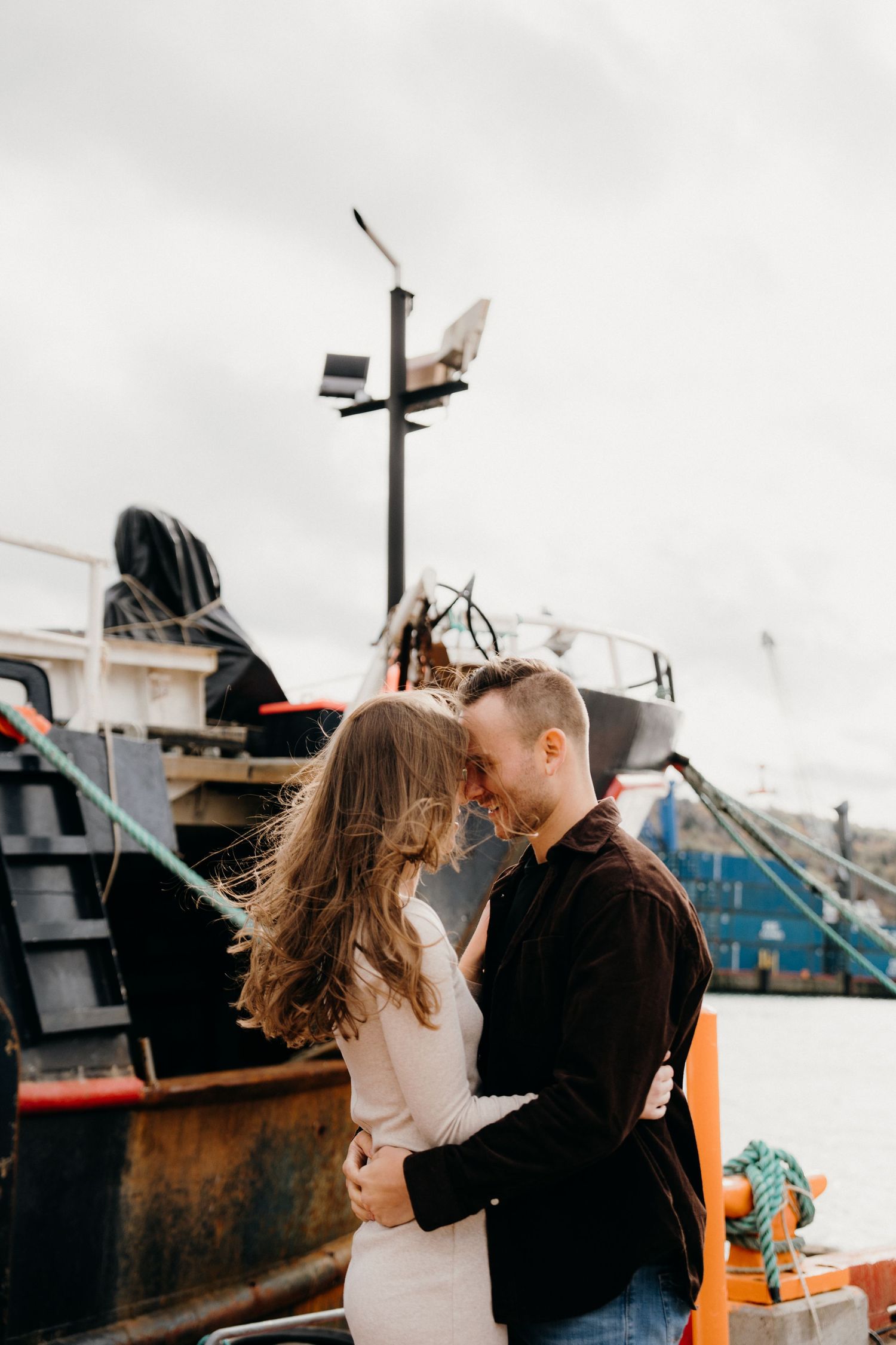 Couple in casual attire shares a tender moment near docked boats on an overcast day.