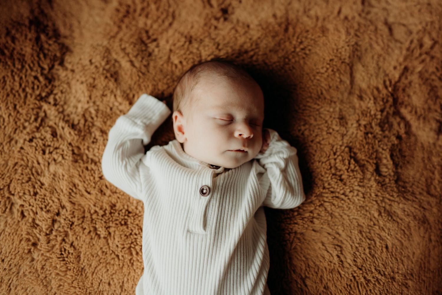Newborn baby wearing white knit clothing resting on textured brown surface.