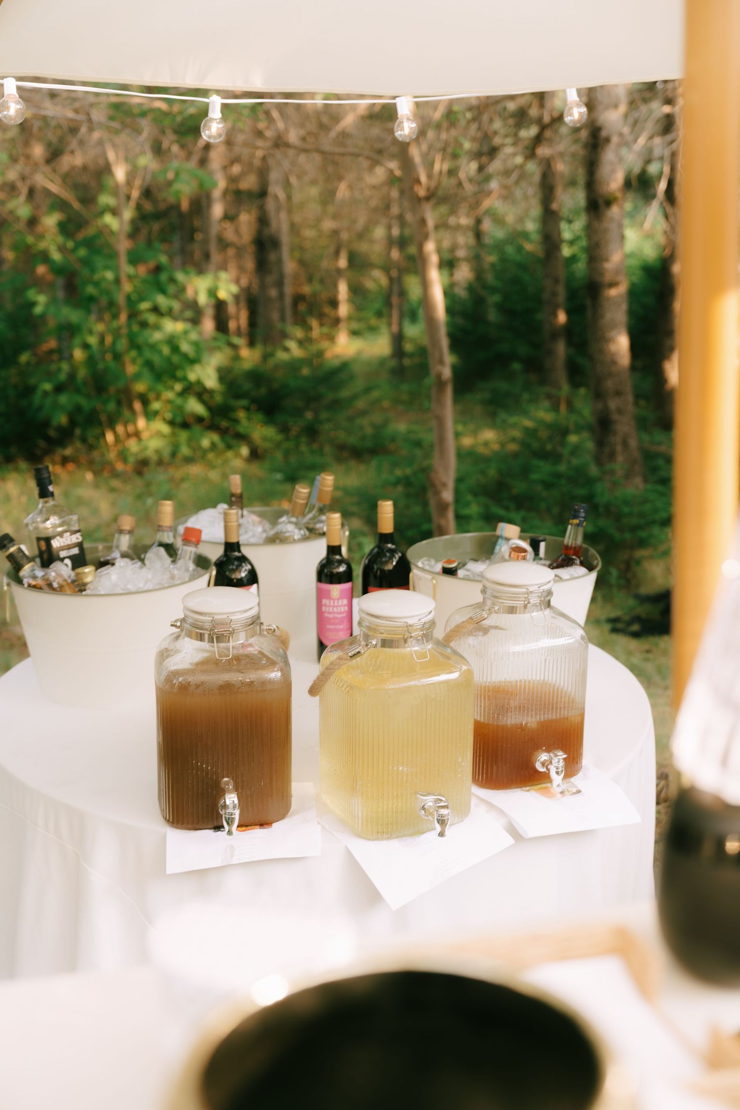 Large glass beverage dispensers filled with various drinks arranged on white tablecloths at an outdoor event.