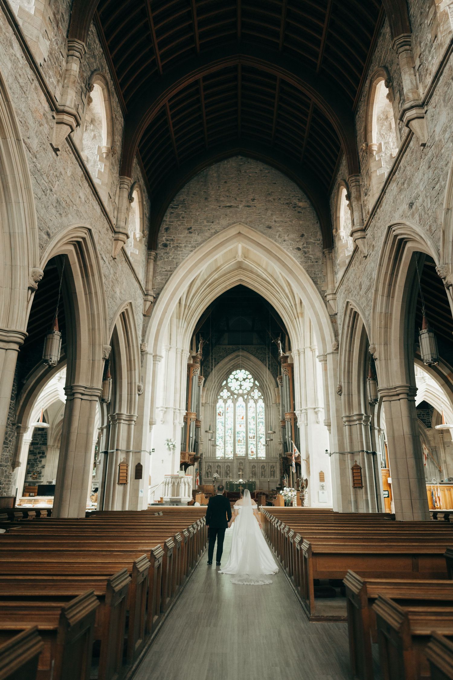 Interior of a gothic church with vaulted ceilings, stone pillars and wooden pews leading to a stained glass window.