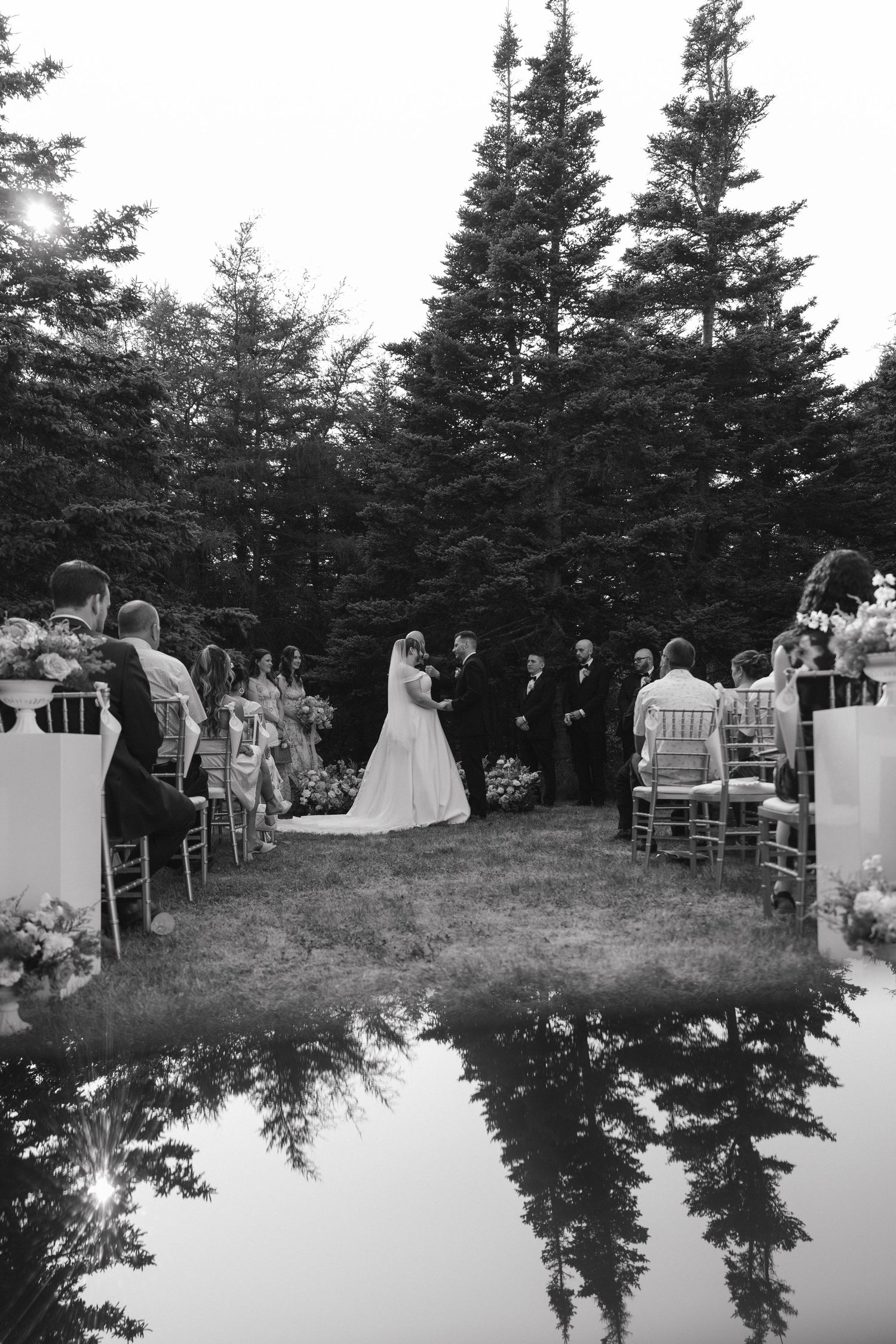 Black and white panoramic view of an outdoor wedding ceremony with pine trees in the background.