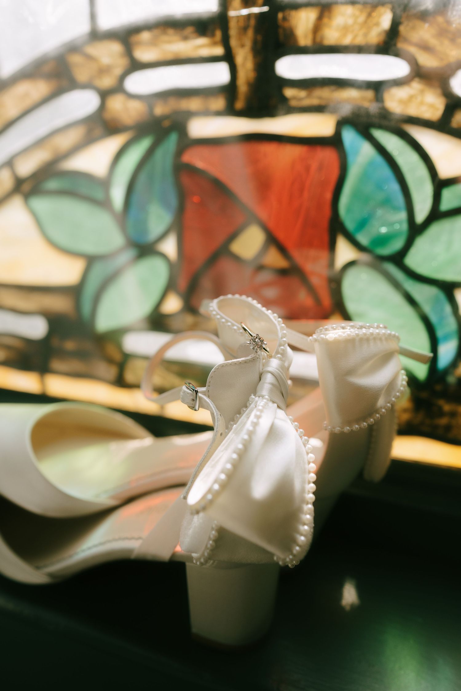White bridal shoes with pearl details photographed against stained glass background.