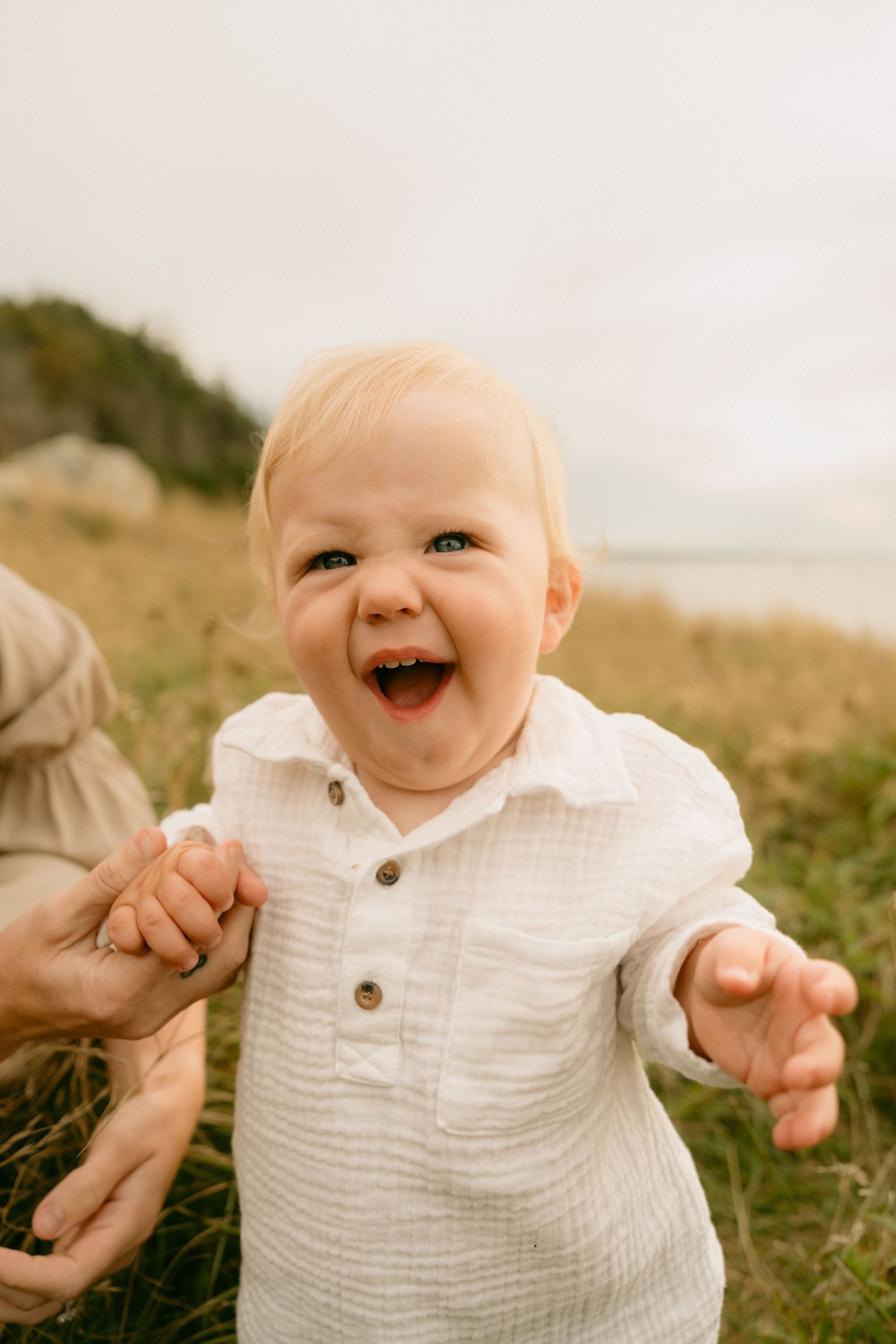 A joyful baby wearing a white muslin shirt laughs while sitting in the grass during an outdoor photo session.