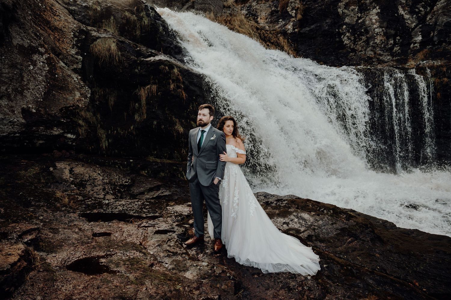 A couple in wedding attire embraces near a cascading waterfall in a romantic outdoor setting.