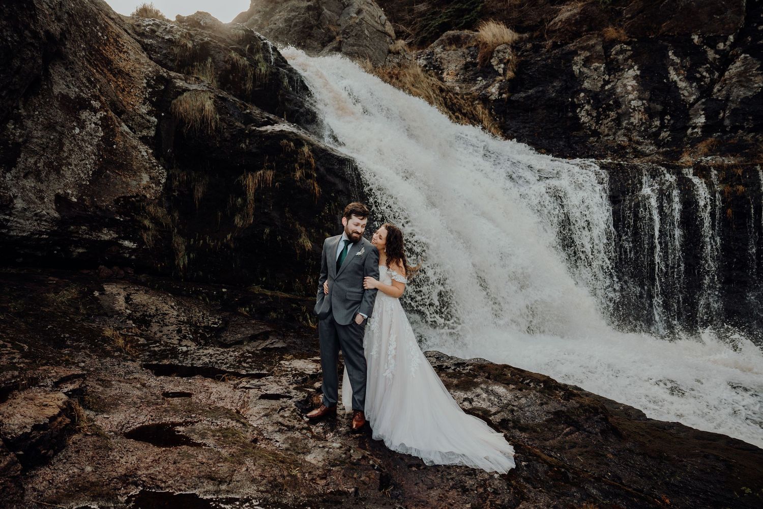 A couple poses near a cascading waterfall in formal attire during a wedding photoshoot.