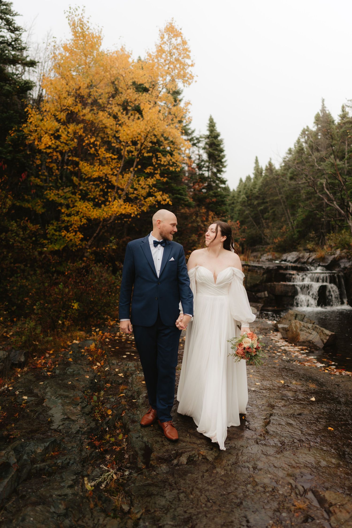 A couple poses together on rocky terrain with autumn foliage and a cascading waterfall in the background.