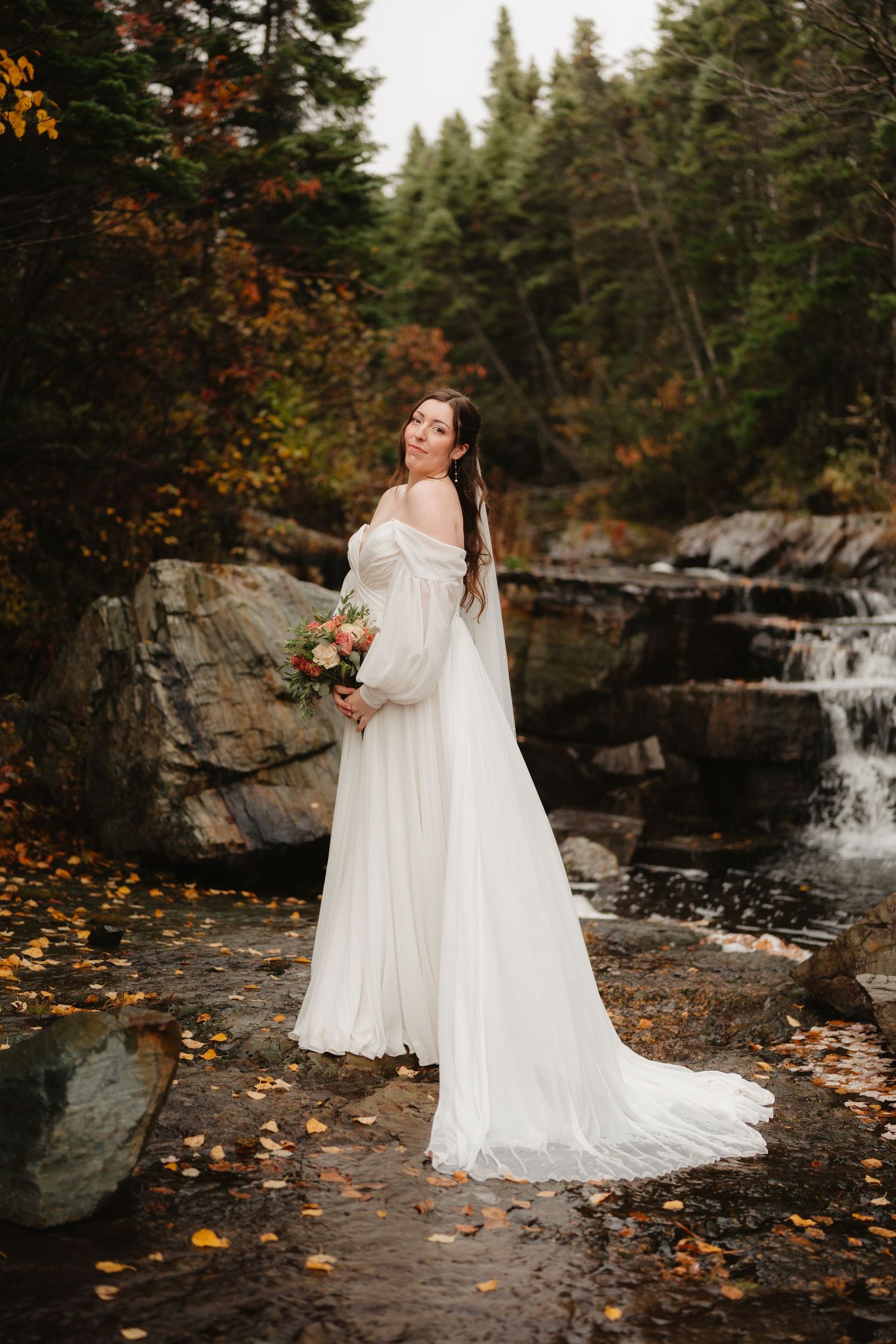 A bride in a flowing white dress poses near a scenic waterfall surrounded by autumn foliage and rocky terrain.