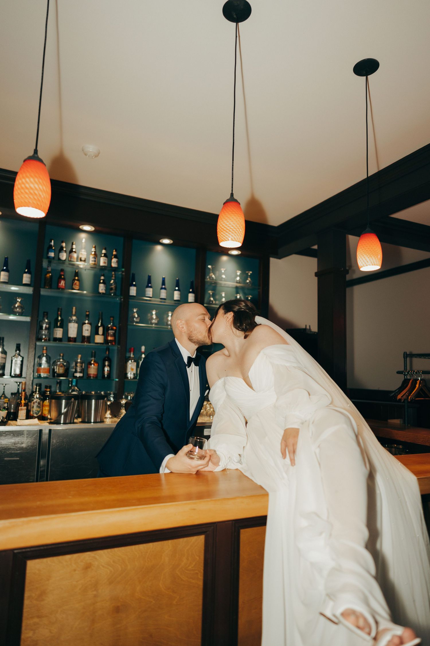 Two people in white shirts lean over a wooden bar counter with orange pendant lights and liquor bottles in the background.