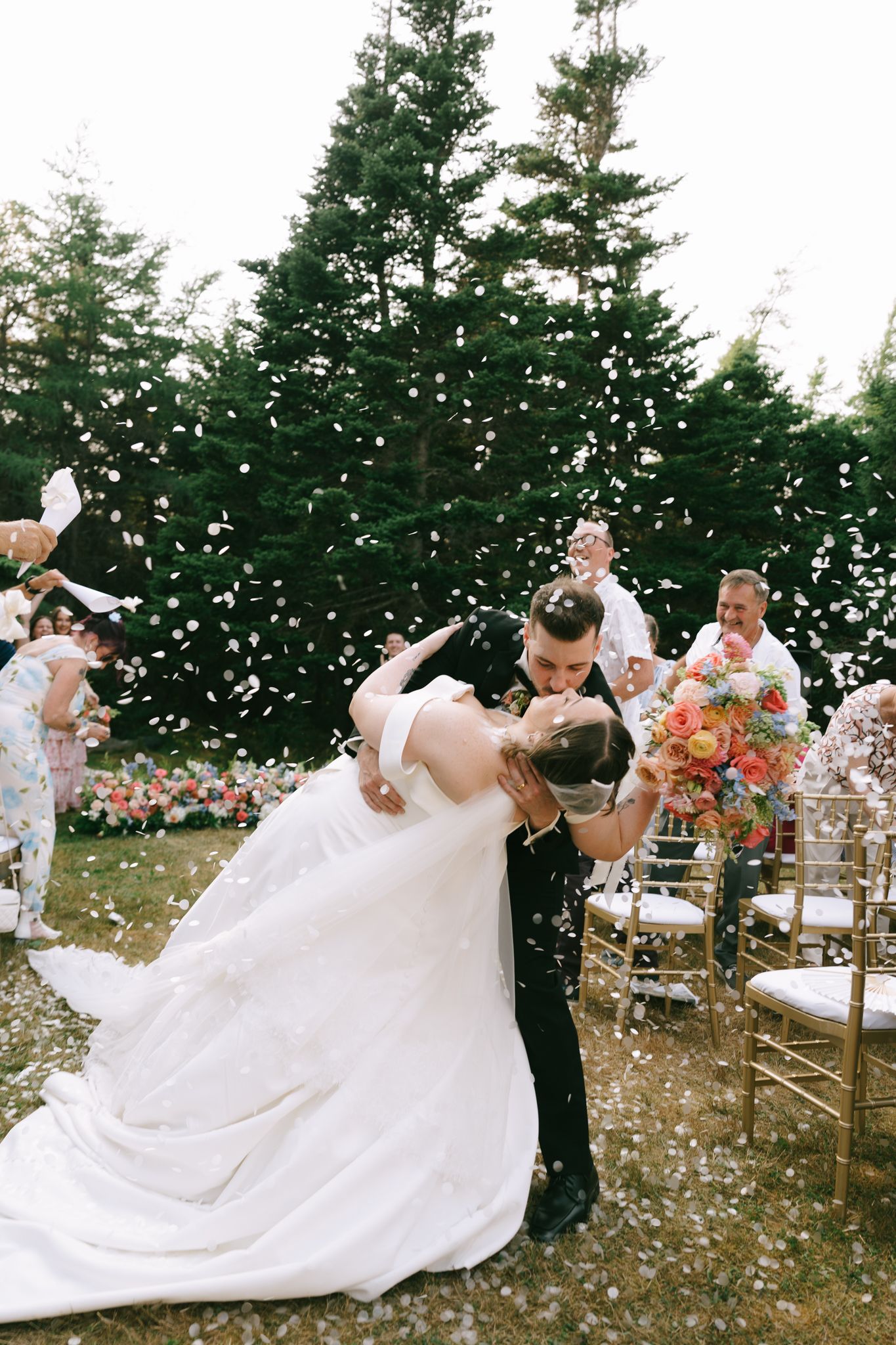 Newlyweds dancing outdoors with confetti floating through the air surrounded by evergreen trees.